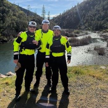 Three firefighters in water rescue gear standing next to river